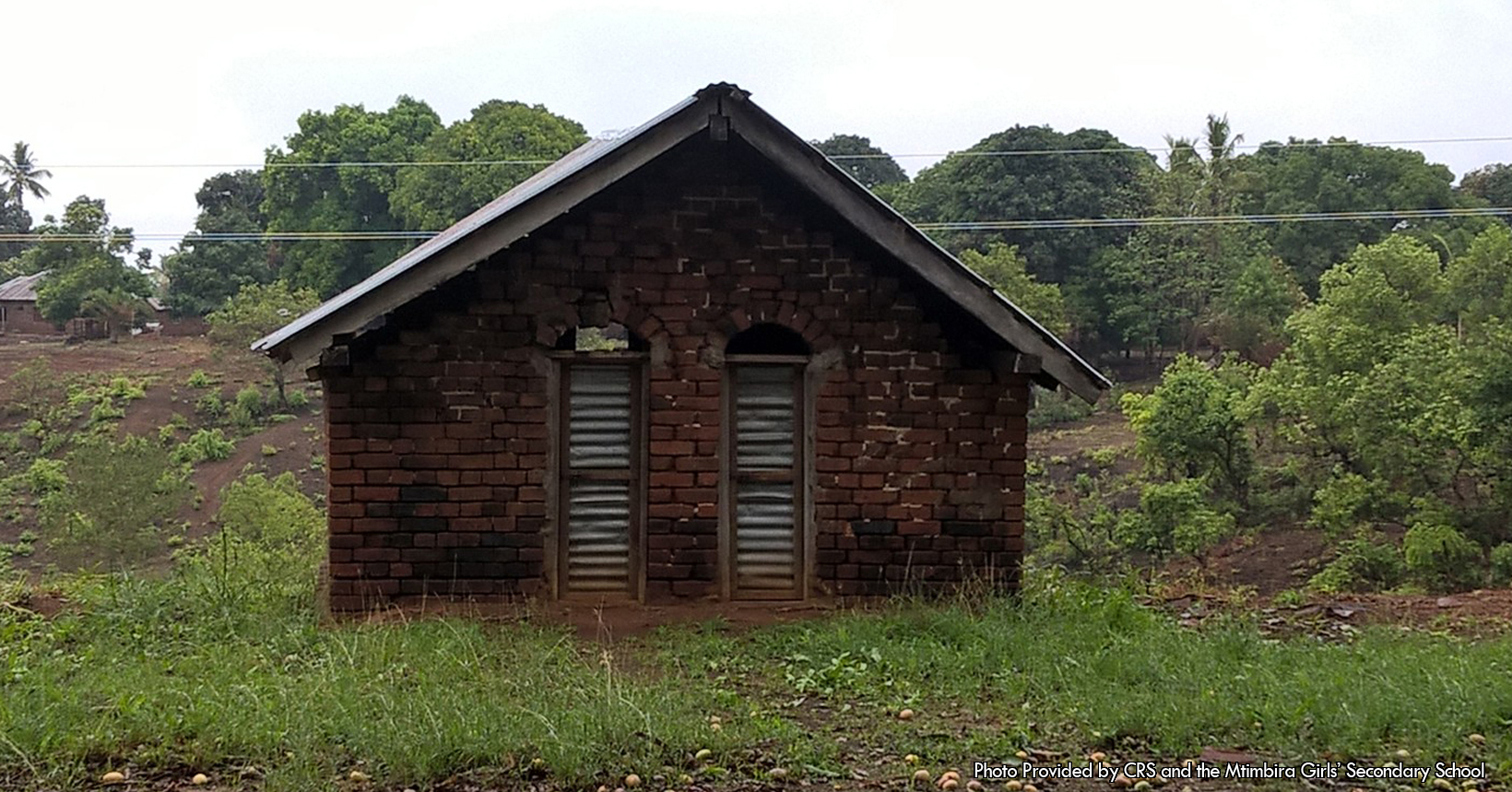 This picture was taken outside of the Mahenge School of Tanzania. This particular building structure is used for toilets. Although when it comes to this particular building, the toilets in here are old. Again, this small building which houses the toilets has the same structural design as the rest of the school. For instance, the buildings which house the library and staff room, the classrooms and the chemistry and biology labs are all made out of bricks. Not only bricks, but every single roof of these buildings are white in color with white shingles. With that being said the building holding the toilet is made out of bricks with a roof covered in white shingles. Furthermore, in the middle of the photo the building has two doors with an arch opening on top of them. The doors are made out of wood beams as well as gray metal coverings. Furthermore, the entire building is resting on top of a dirt field. THE DIRT FIELD IS large and covers up most of the photo. In front of the toilets is a section of the dirt field with a couple of patches of grass laid out. Not to mention there are a few rocks scattered throughout the dirt. Behind the building is the rest of the dirt field. Like in front of the building, this part of the dirt field also has patches of grass scattered around. In addition there are a couple of trees located to the left and right of the photo. Most of the trees to the right are bigger in size and cover up most of the dirt field. Located in the top left corner of the photo is another building on the dirt field. It is also made out of bricks like the rest of the buildings in the school.