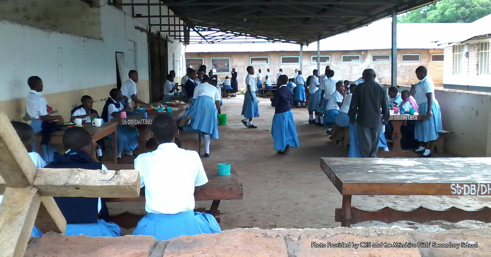 Taken right outside of the school, this photo captures a group of students sitting down and eating. This photo was given to the Fathers Table Foundation by the Catholic Relief Services. The picture was taken inside Tanzania, a country located in East Africa. Here in this photo all of the girls are temporarily down with their studies and are having lunch. Each of the girls have a plate of food as well as a cup to drink. However, some of the girls have different color cups. For instance, one of the girls who is sitting in the left bench has a blue cup while other girls have pink or white cups. Meanwhile, all of the girls who are sitting down or standing up are wearing the same attire. Each of the girls are wearing a light blue dress with a white collared shirt. However, there are some girls who are wearing an extra piece of clothing. In each section of the photo you can see that some girls are wearing sweaters over there white collared shirt. Most of the sweaters are dark blue in color while there is one sweater that is pink, which can be seen on the right side of the photo. Surrounding the girls in their eating area are three buildings, one to the left of the photo and the others are to the right and in the back. The building to the right of the photo is made out of bricks like most of the school buildings are. As for the other two buildings that surround the girls, they are not made out of bricks. Additionally the man in the bottom right corner is one of the many adults that work inside the school. He helps look after the young girls as well as making sure they are well educated.