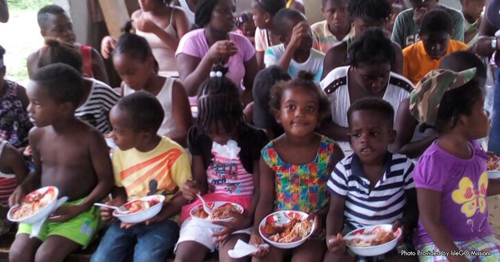 Nutritional hot meals are one of the many things the kids here in the Compound are lacking. Although the Compound lacks a lot of things and has major problems, hunger is one of the major ones. Starting out IsleGo had already been producing nutritional hot lunches out to the children in the second week sincve they have been at the Compound. They are looking to expand this operation to the 4th and 5th week during their stay. Additionally, farm land has already started to be prepared for the Compound for future use. Since there is little to no government assistance as well as assistance from other people living on the island, the people in the Compound will have to learn to survive on their own. This picture which was taken in front of some kids, was given to us by the people from IsleGo Missions. Here there is a group of young children who live in the Compound receiving their own bowl of a nutritional meal. The bowl contains a piece of chicken, rice and other food items to help combat their hunger. IN THE FRONT of the picture is a row of seven kids who are enjoying their meal. In the right side of the picture is one girl who is wearing a purple shirt with a yellow butterfly. Additionally, this young girl is wearing a camouflaged hat and multi colored pants. Sitting next to her is a young boy in a striped shirt. He is holding his spoon in his right hand as he looks off into the distance eating his food. Sitting next to him is another girl who is smiling at the camera. She is wearing a shirt that has squares of man different colors. To her right are four other kids who are also enjoying their meal.