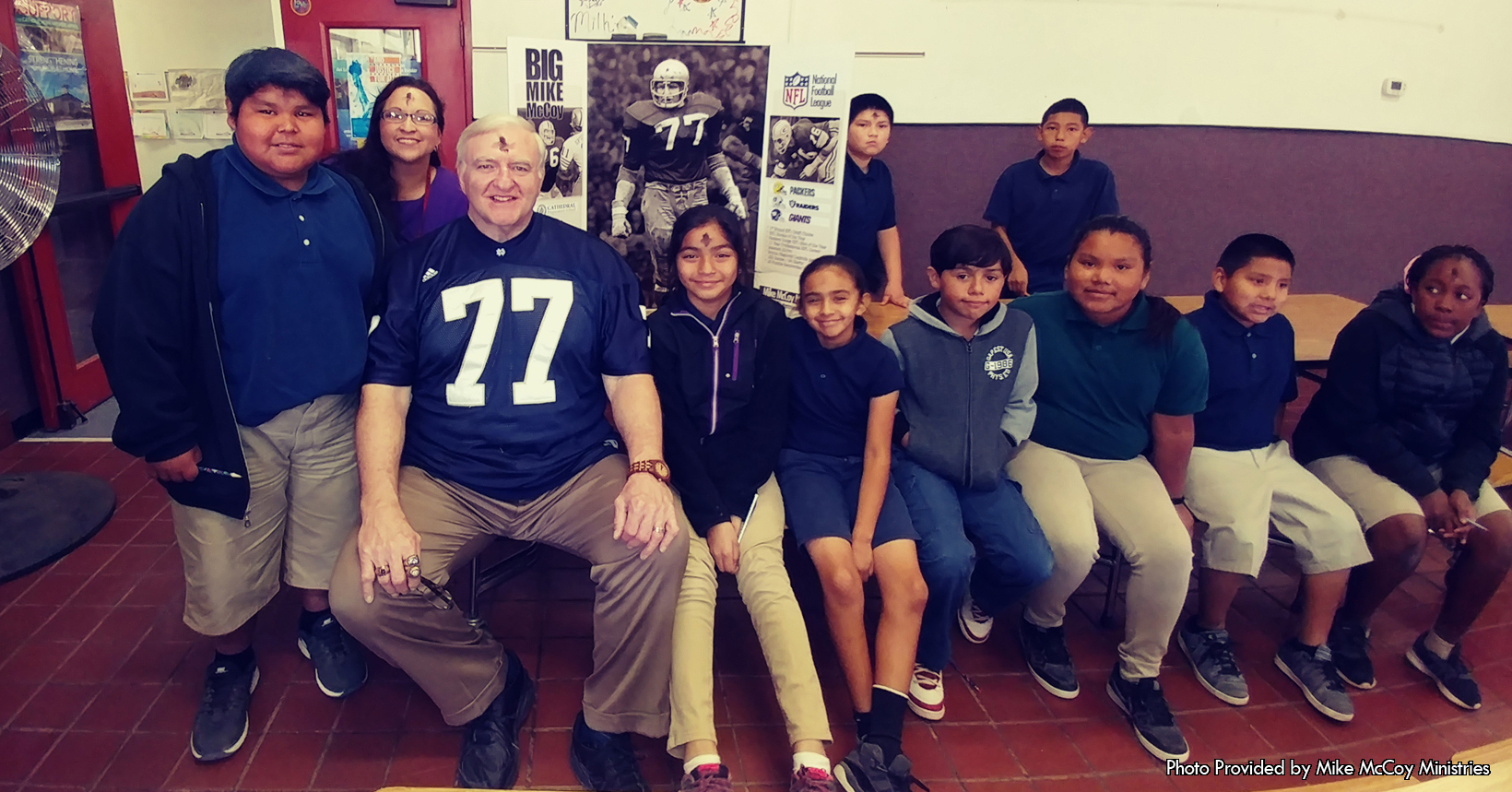 The described photo was taken and provided to us by Mike McCoy Ministry.  To the left of the photo are two young girls. Both of the young girls are wearing white polo shirts. Although the both of the girls are wearing the same style shirt, the young woman on the left has a blue collar while the girl on the right has a standard white collar. Furthermore, both of the girls are wearing skirts that are tan in color.  Around both of their necks are lanyards that are yellow in color and at the end of their lanyards are badges that have their name as well as a photo of them.  To the right of the girls is a trifold presentation board. The board is a representation of Mike McCoy’s life. On the left side of the trifold are few brief descriptions on certain aspects of Mikes Life. On the very top is his name “Big Mike McCoy” in bolded black letters. Under his name is a picture of his younger years when he was playing football going against a player from the opposing team. Furthermore, under this picture are two paragraph descriptions that detail his school years. In the first paragraph, it describes his time where he attended his Cathedral Prep School, as shown by the logo of the school above the paragraph. As for the second paragraph, this one describes his college life. Mike McCoy attended the University of Notre Dame where played football during his college years. You can see the logo of the University of Notre Dame right next to the young girls left wrist. On the right side of the trifold is Mike’s time in the NFL. Mike was selected by the Green Bay Packers as the second overall pick. Besides playing for the Green Bay Packers, Mike also played for the Raiders and the New York Giants. To the right of the trifold is the current day Mike McCoy as he wears a jersey with his number on it, where he has his hand on a young boy from the same school as the two young girls.