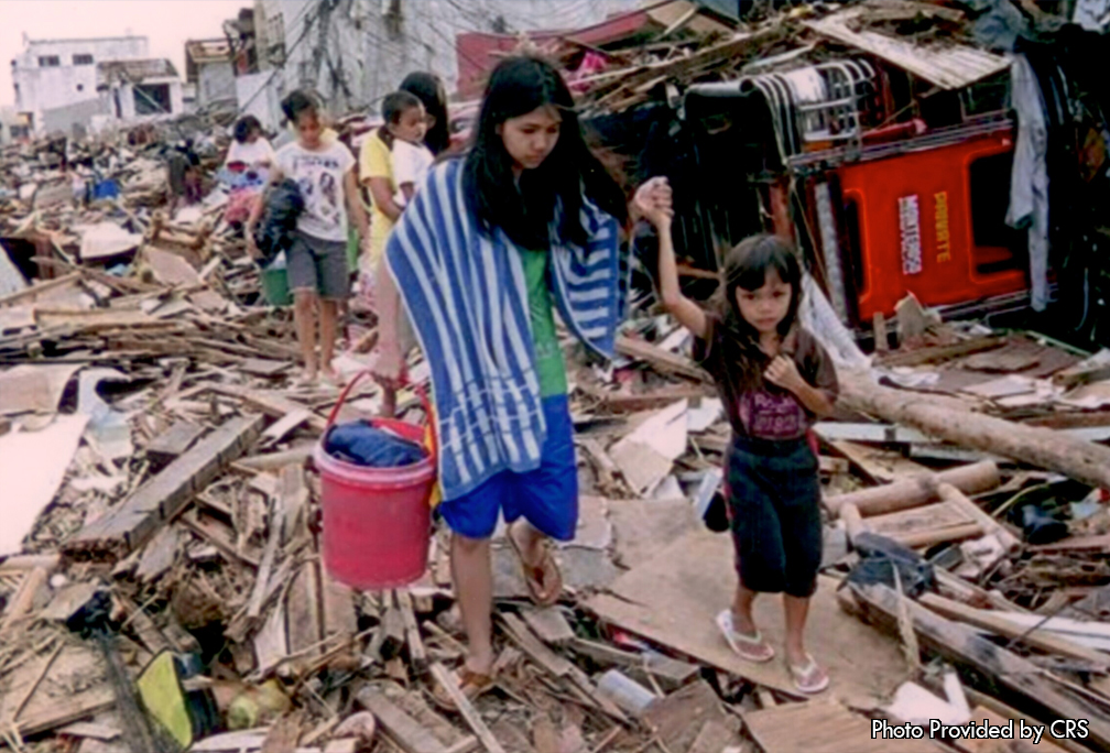 In the photo you can clearly see some of the buildings in the background that are still standing up. The first one you can see is the tall white building in the top left hand corner of the photo.  Most of the building is still standing but there are sections of the building that have been torn off. Towards the right of the building are more structures that have been damaged. Wires have been placed all around them who originally came from phone polls. Furthermore, all around the photo is debris that has come from all over the city. Under the feet of all the people in the photo are multiple pieces of wood that has been scattered. From giant pieces of wood planks to tree branches, there is so much on the ground that you can no longer see the street that use to be there.  In addition, the people in the photo are trying to get to shelter to get out of the damaged city.