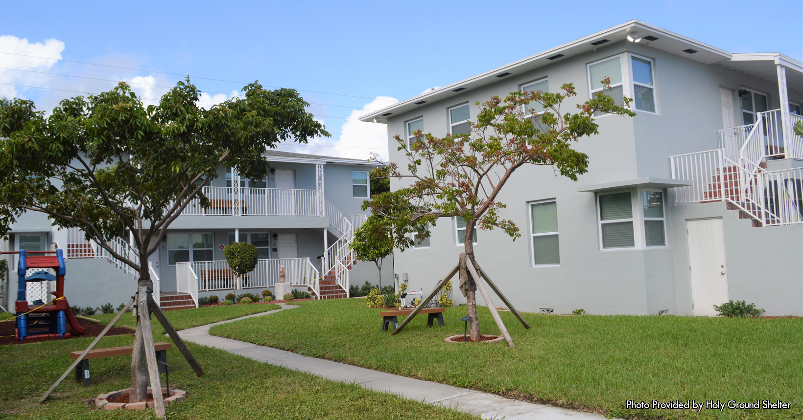 On this beautiful partly cloudy day Phyllis Turner Jepson the executive Director for Holy Ground PBC, took a photo of an apartment complex. This apartment complex was and is used by all of the clients that are associated with the Holy Ground PBC organization. The actually name for the apartment complex is the Stockard Family Campus. This apartment complex was purchased by the Holy Ground PBC organization back in November of 2017. The Holy Ground PBC organization then renovated the apartment complex in 2018. Here at the Stockard Family campus they hold many events for the organization and the clients. As a matter of fact, on May 3, 2018, the Holy Ground PBC held their Ribbon Cutting Ceremony. Going into more detail, the Stockard Family Campus is made up of many different sections. The campus is made up of apartments for the clients, housing for the Resident Advisor, a work space for staff members and a community suite. The community suite is used by clients of the organization for community meetings and celebrations. Furthermore, the community suite allows clients to study together, use the computer stations and even to watch television and movies together. Having their very own apartment complex allows the staff members and the clients to have a better sense of a community. It has been a true blessing as it also allows Holy Ground to change the lives of its clients for the better. In the photo taken by Phyllis you can see two buildings that are a part of the Stockard Family campus. Both of the buildings are painted in a light grey color with multiple windows on each of its sides. Not to mention both of the buildings are two stories. Each has a set of staircases with white railing that safeguards all who walk. Furthermore, in the bottom left hand of the photo is a small playground that allows the children to have fun and play.