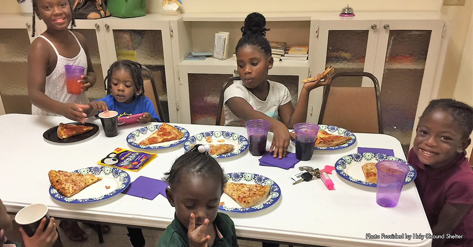 The following photo was taken by Phyllis Turner Jepson who is the Holy Ground PBC’s Executive Director. The following photo was taken inside one of the rooms of the Holy Ground Shelter. Currently during the time of the photo there are several kids in the room. The children in the photo are having a small pizza party. The staff members at the Holy Ground PBC know that the newly young mothers can’t always help their children in their day to day needs. This is very true when it comes to feeding their kids while they are attending their group and individual sessions. This is the reason why the kids are enjoying slices of pizza. In the photo there are 6 kids who are sitting down. Starting from the right side of the photo is the first child eating her slice of pizza. She is wearing a maroon dress that goes all the way down to her ankles. Sitting in front of her on the table is her own slice of pizza. Like some of the other young children sitting down with her this young lady is eating a cheese pizza. Next to her pizza is her purple cup that is empty since she drank all of her soda. To her right are three more young children who are also enjoying their slices of pizza. All three of them are eating a slice of pepperoni pizza, while two of them are drinking orange soda and the other is drinking coke. On the opposite side of the table there are the last two young kids who are eating there slices of cheese pizza. These two kids are both wearing a green shirt and white cargo pants. The Holy Ground PBC has nannies that have undergone background checks and are trained in childcare. The nannies make sure that the parents don’t have to worry about their kids and attending their sessions.