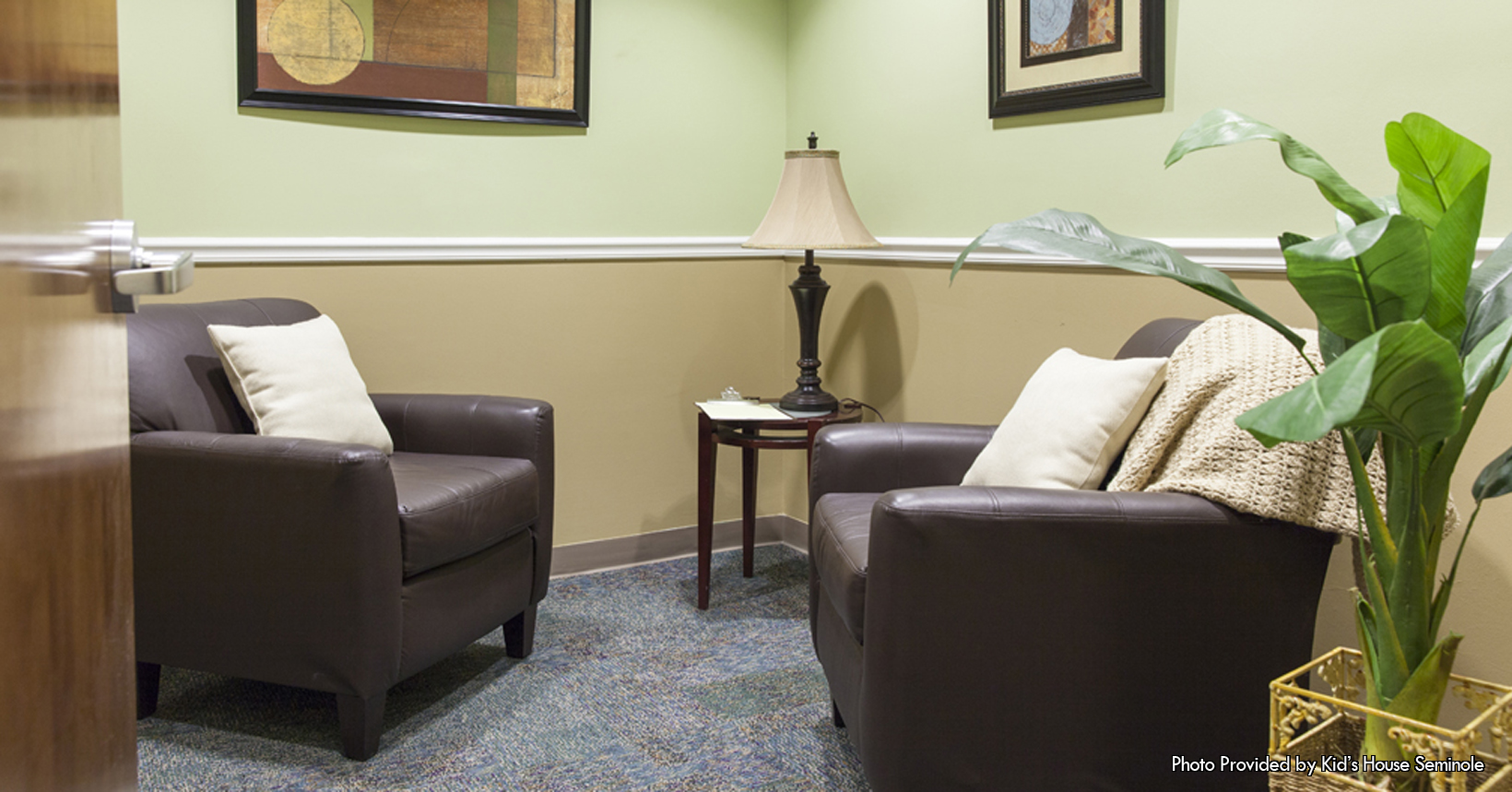 The following picture was taken inside one of the rooms located in the Kids House of Seminole and was provided by them as well. The office is painted in a tannish color and has many different objects mounted on the walls. Firstly, in the left side of the picture there are two things mounted on the wall. The first thing mounted on the wall is a picture of a tiger that is in between two plants. Directly to the right of the picture on the wall is a standard TV mounted on the wall. On the other hand, there is only one thing on the other side of the wall. Here there are only two picture frames with one document framed in each of them. In addition to things being on the wall of the office, there are also things in the middle of the office. Starting in the back of the office, there is an office phone used to communicate between members in different rooms in the Kids House of Seminole building. As well as the telephone there is a table in the middle of the office. This table is shaped like an oval with a red trim that goes around the entire table. Additionally, there is a box of Kleenex tissue that is sitting on top of the table in the office. Surrounding the table is a group of six chairs. These chairs are black in color and are made from leather. Coupled with being made in leather, the arm rest of the chairs have leather cushions on them to help ease the pain of the arms of those who sit in the chair. This room is used to hold meetings to decide the next step for the Kids House of Seminole, and to make sure that kids in the area have a safe place to stay during their hard times of being abused.