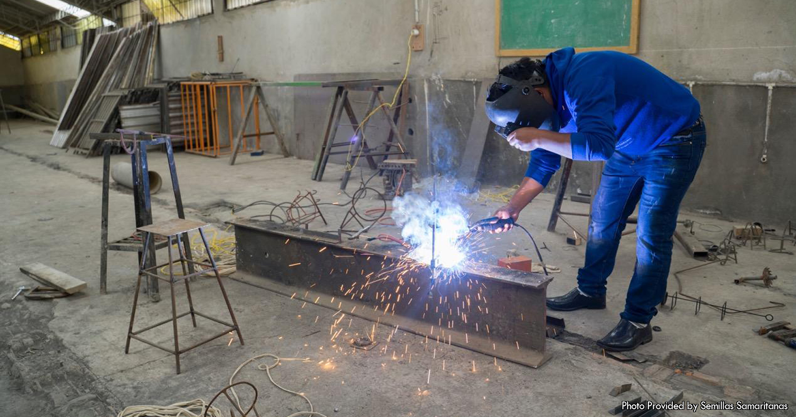 This picture was taken by the Semillas Samaritanas project and was given to the foundation. This photo was taken inside of the metal working workshop. Here in the workshop, the doors and the windows that the Semillas Samaritanas project requires are built. In addition to being the place where the doors and windows are produced, this workshop has also been used to make other things for different people. Meaning that other people outside of the Semillas Samaritanas project have requested things to be made in the workshop. In the middle of the photo is one of the people who works inside the workshop. During the time of the photo the young man is wearing a dark blue hoodie. In addition, the young man is wearing blue jeans with black slip on shoes. During the time the photo was taken, the young man is in the metal workshop welding a steel beam. His blow torch is heating up the piece of steel to allow the beam to connect to something. In addition, the young man in the workshop is wearing a protective mask to keep the sparks from flying into his eyes and face. Located all around the young man and the workshop are various items used in the workshop. For starters, there is a small stool in front of the steel beam. Not to mention, there are multiple pieces of metal that are sitting on the floor of the workshop. In addition, there are multiple wires that are also laying on the floor. In the bottom left corner of the picture, there is a long white wire that is coiled up on the floor of the workshop. Not to mention, there are a couple of additional objects in the back of the photo that are also used in the workshop by the