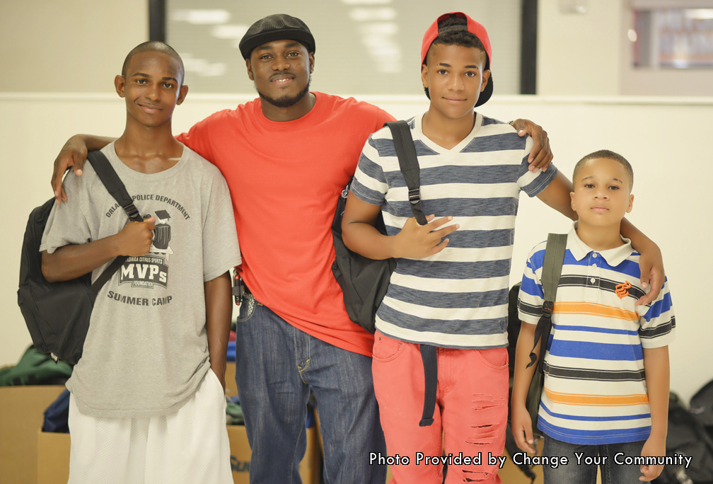 This photo was taken inside the building where backpacks and school supplies were being handed out. In addition, this photo was taken by the staff member of the Change Your Community organization. After the photo was taken and the even was over the staff members sent this to the Father’s Table Foundation. Looking at the photo there are four men standing and posing. Three of the men in the photo are young kids who come from low-income families that don’t have access to a lot of supplies. For that very reason they came to one of the annual Give-A-Ways to get some school supplies. Starting from the left of the photo is the first young boy. He is wearing a gray t-shirt that came from the Orlando police department. On the shirt is a stick figure that has a cap and gown and is graduating. Furthermore, he is wearing white basketball shorts as he smiles and wears his backpack with his new school supplies. Next to him in the red shirt is a staff member for the Change Your Community organization. He is here supporting the young kids who are here to get school supplies and occasionally sneakers. He is smiling because he knows that the kids who are coming are going to leave better than when they came. Next to him is the second young man. He is wearing a red snap back hat as well as a stripped t-shirt. In addition, this young man has red long pants as he wears his new backpack on his back. Finally to the far right of the group is the youngest of the kids. This young man is wearing a collared shirt that has white, blue, orange and black stripes.